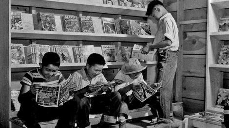 Four youth — confined to the War Relocation Authority center in Newel, Calif. — enjoy the latest comic books available at the newsstand in July 1942. In the upper right, issues of "Fantastic Adventures" (August 1942) and "Wild West Weekly" (June 20, 1942) await sale.