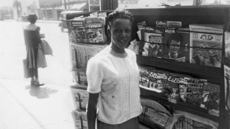 A woman poses in front of a street-side newsstand in fall 1943. The stand carries "Doc Savage" (September 1943), "The Masked Detective" (Spring 1943) and "Exciting Love" (Fall 1943).