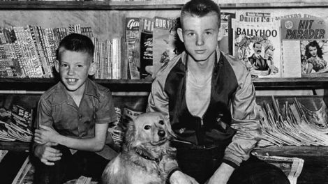 Two boys pose in front of a magazine stand in the summer of 1948. A number of pulp spines can be seen on the shelf at left.